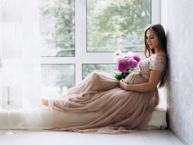 Sensetive portrait of pregnant woman. Expecting lady lies on the windowsill in a bright room