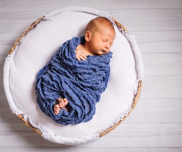 Newborn baby enveloped in blue scarf sleeps on white pillow