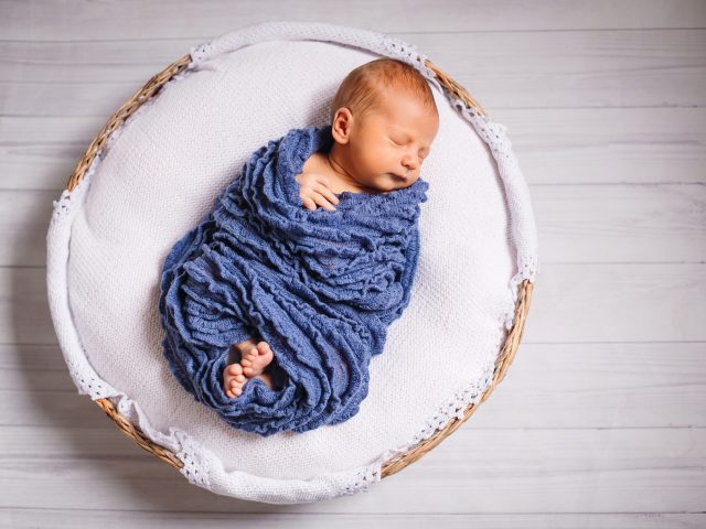 Newborn baby enveloped in blue scarf sleeps on white pillow