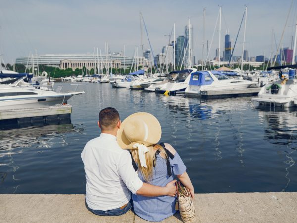A beautiful and elegant woman with light hair and a blue dress sitting in the summer solar park near water with her husband dressed in a white shirt