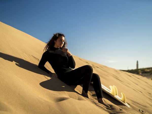 A shallow focus shot of an attractive female posing on a sandy hill with a surfboard on the side
