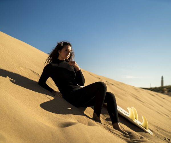 A shallow focus shot of an attractive female posing on a sandy hill with a surfboard on the side