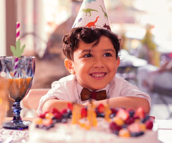 Adorable birthday boy. Dark haired optimistic boy wearing party hat with dinosaurs and smiling while sitting with his birthday cake