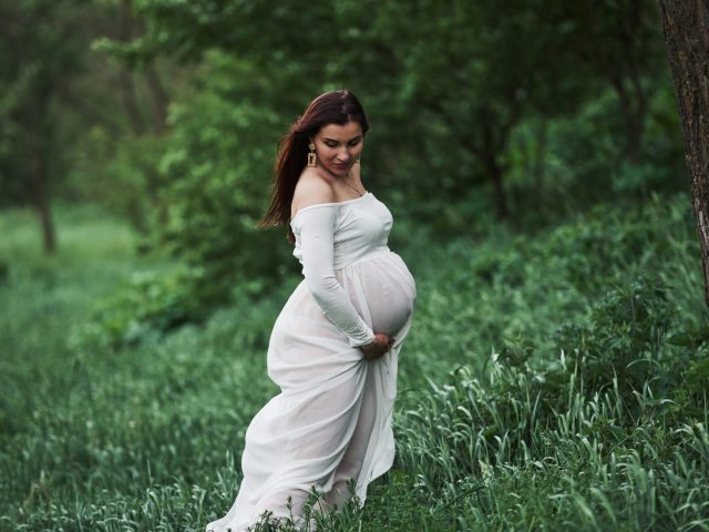 Standing in the grass. Beautiful pregnant woman in dress have a walk outdoors. Positive brunette.